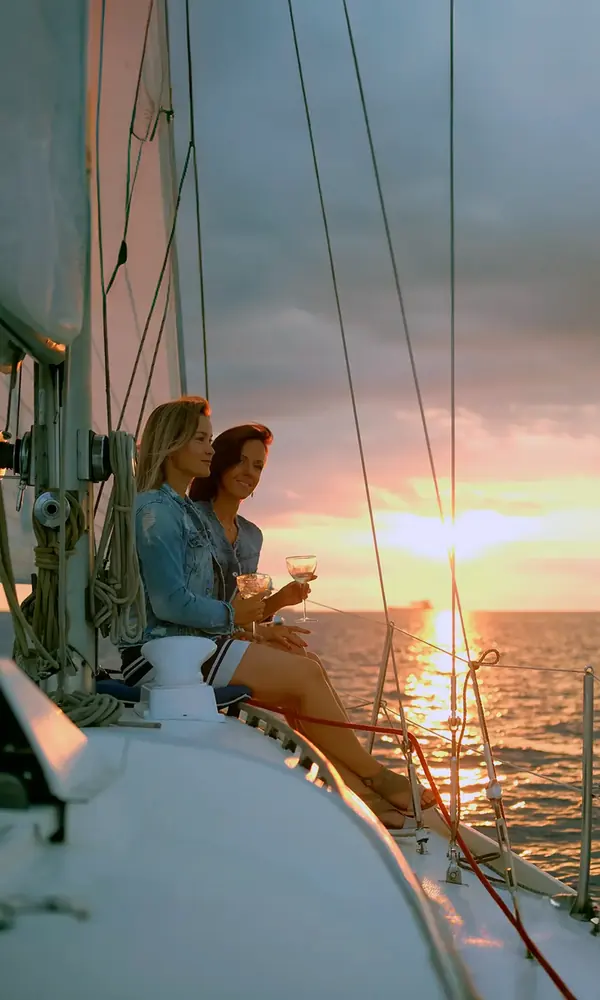 Two guests on the bow of a sailboat with a glass in hand, watching the sun touch the horizon